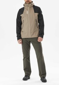 Beige und schwarze Jacke mit Zipper, zwei seitlichen Taschen und Kapuze. Kombiniert mit olivgrünen Hosen und schwarzen Schuhen.