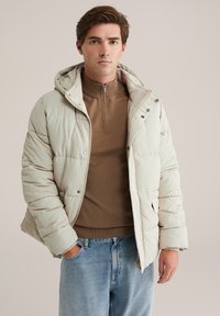 Chaqueta tipo plumón en beige claro con cierre frontal y capucha. Presenta un diseño acolchado, bolsillos laterales y se lleva sobre un sudadera marrón con cremallera.