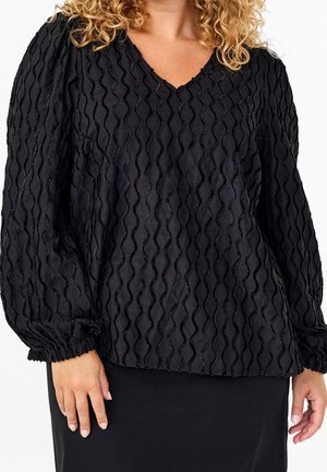 Curly-haired person wearing a textured black long-sleeve V-neck blouse with gathered cuffs and a black skirt.