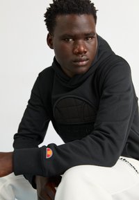 Sudadera negra con diseño acolchado en el pecho, ajuste cómodo y logo bordado en la manga. Modelo sentado contra un fondo neutro.