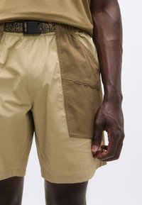 Μπεζ σορτς με αντίθετη καφέ πλαϊνή τσέπη, κατασκευασμένα από ελαφρύ υλικό. Διαθέτουν ελαστική μέση και ζώνη με σχέδιο.