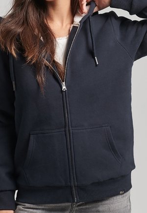 Frau trägt einen dunkelblauen Kapuzenpullover mit Reißverschluss und Vordertaschen sowie ein weißes Hemd darunter, teilweise geöffnet, Hand richtet die Kapuze.