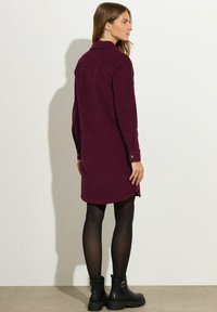 Kastanienbraunes Cordhemdkleid mit langen Ärmeln, Knopfmanschetten und abgerundetem Saum. Kombiniert mit schwarzen Stiefeletten und transparenten schwarzen Strumpfhosen.