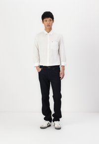 Witte lange mouwen overhemd met knopen en een zak, gecombineerd met donkerblauwe jeans en grijze sneakers; eenvoudig ontwerp en fitted silhouet.