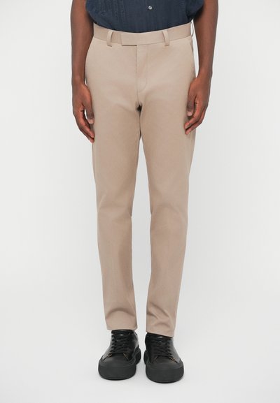 Beige maßgeschneiderte Hose mit flachem Frontschnitt, Gürtelschlaufen und schlanker Passform, kombiniert mit schwarzen Sneakers. Glatte Textur und klare Designelemente.