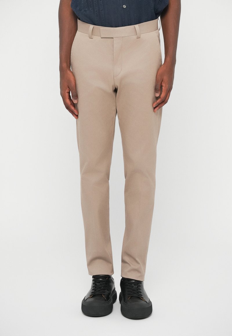 Μπεζ παντελόνια κομμένα με επίπεδο μπροστά, τσέπες ζωνάκι και στενή εφαρμογή, φορεμένα με μαύρα αθλητικά παπούτσια. Λείος υφασμάτινος χαρακτήρας και καθαρές σχεδιαστικές λεπτομέρειες.