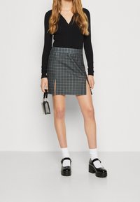 Svart långärmad topp kombinerad med en knälång grå kjol med rutmönster och en sidoflik. Modellen håller en liten svart handväska.