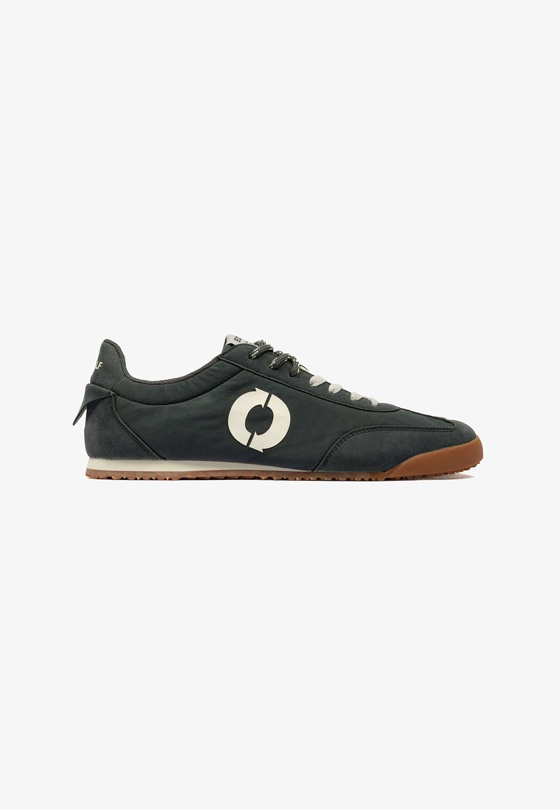Baskets noires à profil bas avec logo flèche circulaire blanc, lacets gris et noirs, semelle en caoutchouc marron avec grip texturé.
