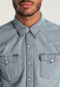 Ljusblå denimskjorta med spetsig krage, silverfärgade tryckknappar och två bröstfickor. Har en texturerad yta och västerninspirerad yoke.