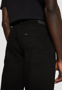 Hombre vistiendo jeans negros y una camiseta negra, mostrando los detalles de los bolsillos traseros y la cintura de los jeans.