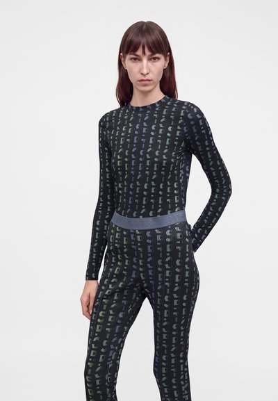 Femme aux cheveux bruns raides portant un haut noir ajusté à manches longues et un pantalon assorti avec un motif à rayures verticales.
