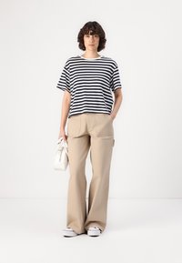 Tricou oversized cu dungi albe și bleumarin, pantaloni largi bej, adidași din pânză, ținând o geantă mică albă. Textură de țesătură netedă.