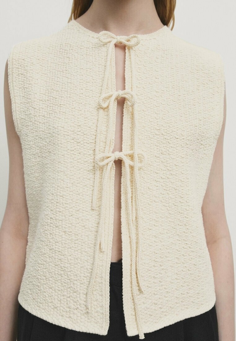 Gilet sans manches crème texturé avec trois nœuds noués sur le devant, porté ouvert sur un pantalon noir.