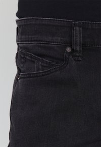 Acercamiento del área del bolsillo de unos jeans de mezclilla negra con el lazo del cinturón visible y remaches de cobre sobre un fondo de tela oscura.