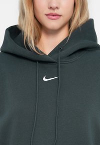 Tamsiai žalia pūkinė striukė, pagaminta iš minkštos medvilnės mišinio, su balta Nike logotipu ant krūtinės, raišteliais ir dideliu gobtuvu.