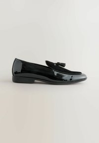 Schwarze Lackleder-Slipper mit flachem Absatz, ausgestattet mit einem Wildlederakzent auf dem Obermaterial und einem dekorativen Quasten-Detail. Glatte, glänzende Oberfläche.