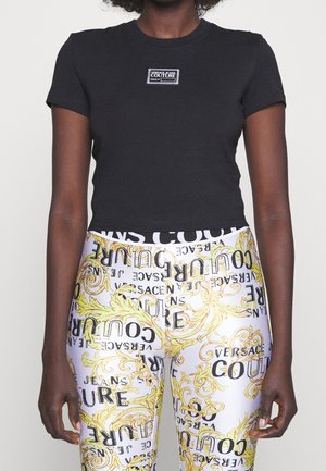 Kobieta w czarnym krótkim topie z logo Versace Jeans Couture i białych legginsach z żółtym barokowym wzorem i tekstem marki.
