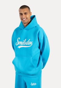 Sudadera azul de suave tejido con un bolsillo frontal y un gran logo blanco de "Smilodox". Combinada con pantalones azules a juego.