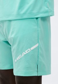 Ένα άτομο που φοράει αθλητικά σορτς σε ανοιχτό τιρκουάζ και ένα αντίστοιχο τοπ, με το λευκό λογότυπο "HEAD" στα σορτς.