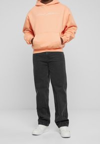 Persona che indossa un hoodie pesca con una scritta bianca, jeans neri e sneaker bianche, in piedi su uno sfondo grigio chiaro uniforme.