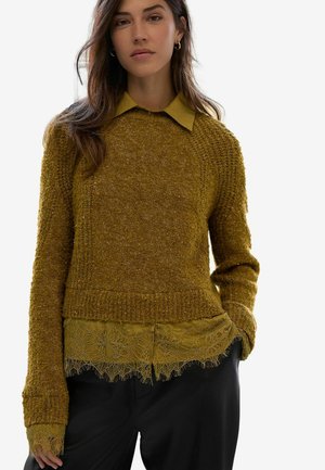 Senfgelber strukturierter Strickpullover, der über einem mit Spitze besetzten Hemd getragen wird, mit Kragen und langen Ärmeln mit Rippbündchen. Schwarze Hose.