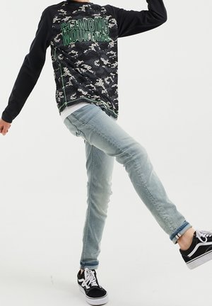 Schwarzes langärmeliges Oberteil mit Camouflage-Muster und grünem Text "Climbing Mountains", kombiniert mit hellblauen Jeans und schwarzen Turnschuhen.