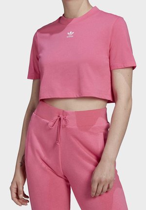 Osoba nosící růžové crop top tričko Adidas a odpovídající kalhoty se stahovací šňůrkou na jednobarevném pozadí.