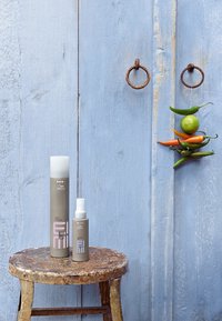 Two Wella hair products on a rustic stool in front of a weathered blue wooden door with rusty metal rings and hanging chili peppers and lime.