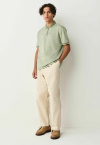 Tricou polo cu model verde, cu guler și detalii albe, purtat cu pantaloni bej și pantofi din ante maronii.