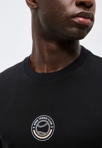 Gros plan d'une personne portant un T-shirt noir avec un logo "Boss Tennis Club 1997" comportant un graphique de balle de tennis.