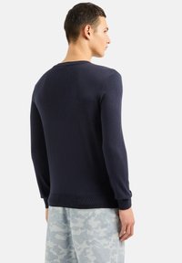 Maglione a maglia blu navy con maniche lunghe, scollo rotondo e polsini a coste. Indossato con pantaloni grigi chiari a motivo camouflage.