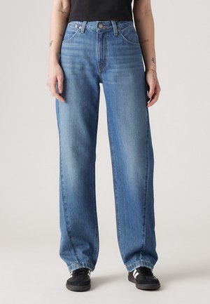 Personne portant un jean bleu clair à jambes droites, des baskets noires avec des bandes blanches et un haut noir, debout devant un fond uni.