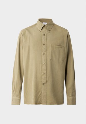 Jeune homme debout portant une chemise beige à boutons, un pantalon noir et des chaussures noires devant un fond blanc uni.