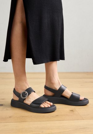 Femme portant des sandales noires à bout ouvert avec des lanières épaisses et une boucle ronde, debout sur un sol en bois, vêtue d'une jupe noire côtelée avec une fente latérale.
