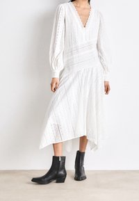 Abito bianco in pizzo con maniche lunghe a palloncino, scollo a V e orlo irregolare. Abbinato a stivaletti neri alla caviglia con cerniere laterali e tacco leggermente curvo.