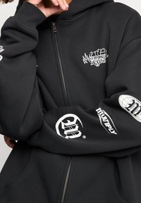 Sweat à capuche noir à fermeture éclair, fabriqué en tissu mélangé en coton, avec des logos blancs sur les manches et la poitrine, des poignets côtelés et une poche avant.