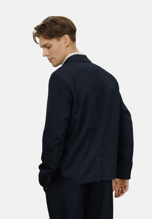 Ung mann med brunt hår iført en tettsittende mørk marineblå dressjakke, som ser nedover og litt til siden mot en ensfarget lys bakgrunn.