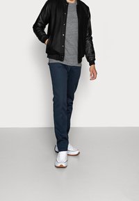 Mand iført en sort bomberjakke, grå t-shirt, blå bukser og hvide sneakers går mod en ensfarvet lys baggrund.