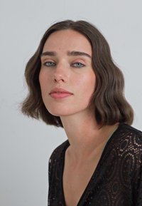 Persona con capelli ondulati di media lunghezza, che indossa un top in pizzo nero con scollatura a V. Uno sfondo chiaro mette in risalto i tratti del viso e l'espressione.