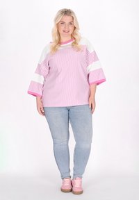 Randig oversized skjorta i rosa och vitt med vida ärmar, kombinerad med ljusblå jeans och rosa sneakers på en modell mot en vit bakgrund.