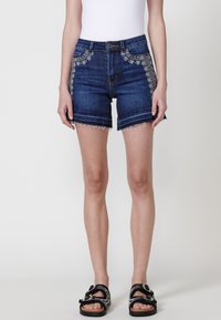 Shorts en denim bleu foncé avec broderie florale sur le côté, ourlet effiloché et coupe ajustée. Associés à des sandales noires avec des lanières ornées.