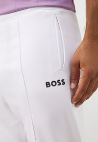 Witte sportbroek met een gestructureerd oppervlak. Heeft een zijzak en een zwart "BOSS" logo op de dij. Hand rustend op de stof.