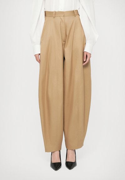 Donna che indossa pantaloni beige a vita alta e gamba larga, camicia bianca a maniche lunghe infilata dentro, e tacchi a punta neri, in piedi di fronte a uno sfondo semplice.