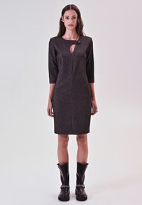 Figurbetontes, dunkelgraues Kleid mit vertikalen Streifen, 3/4-Ärmeln, Wasserausschnitt und einem dekorativen Knopf. Kombiniert mit kniehohen schwarzen Stiefeln.