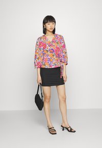 Blusa floral em tons vibrantes de rosa, laranja e roxo; combinada com uma saia mini preta. A modelo segura uma mala preta de ombro, usando sapatos de salto com tiras.