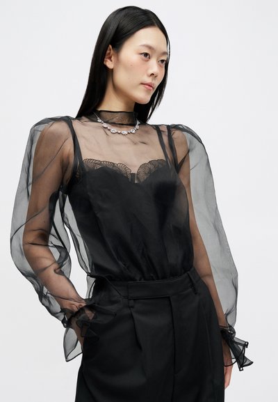 Μαύρη διαφανής μπλούζα με φουσκωτά μανίκια, δαντελένιες λεπτομέρειες και βολάν στους καρπούς, συνδυασμένη με κομμένα μαύρα παντελόνια. Τα κοσμήματα διαθέτουν ανθισμένο σχέδιο.