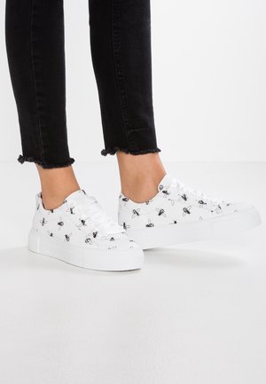 Hvide platform-sneakers med sorte bi-print, båret med korte sorte jeans med frynser på en lys baggrund.