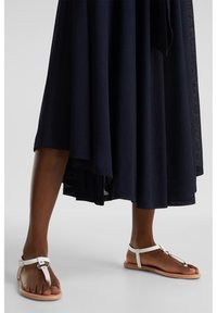 Jambes portant des sandales plates blanches à bride en T avec boucle sur semelles couleur cuir, associées à une jupe midi fluide bleu marine foncé.