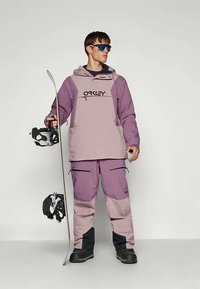 Muž v purpurovém a růžovém oblečení na snowboard drží snowboard a helmu, má na sobě černé boty a brýle se modrým zabarvením.