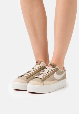 Personne portant des baskets Nike beige avec des logos swoosh blancs et des semelles blanches épaisses, debout sur une surface blanche.